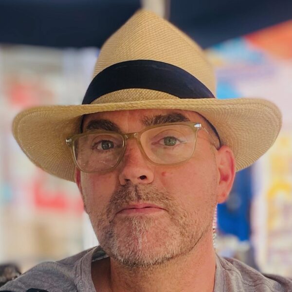 Frédéric, founder and CEO of Wine Tours in Style, wearing a Panama hat and glasses, seated at an outdoor café, exuding a relaxed yet refined presence in an urban setting.